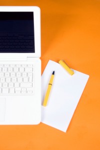 A white laptop, a pad of paper and a yellow pen on an orange background